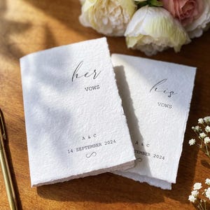 May include: Two white handmade paper vow booklets with deckled edges. One booklet is printed with the text 'her VOWS', 'A & C', '14 SEPTEMBER 2024', and an infinity symbol. The other is printed with 'his VOWS', 'A & C', and '14 SEPTEMBER 2024'.
