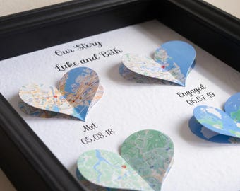 May include: A framed wall art piece featuring three dimensional heart shapes cut from maps. The text reads Our Story, Luke and Beth, Met 05.08.18, Engaged 06.07.19, and Married. Each heart is placed above a date and event label on a white background.