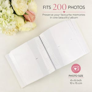 May include: An open white photo album with clear plastic sleeves for 200 photos. The text reads FITS 200 PHOTOS and PHOTO SIZE 10 x 15 cm. A bouquet of cream and pink flowers is positioned in the upper left corner of the frame.