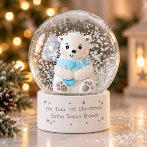 May include: A clear glass snow globe containing a white polar bear figurine wearing a light blue scarf. The base is white with the text On Your 1st Christmas Esme Susan Brown surrounded by small star icons. The globe is filled with white snow particles.