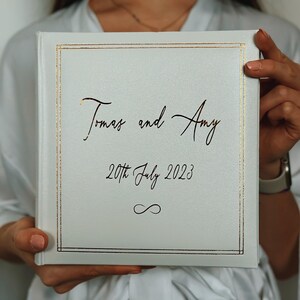 May include: A square white photo album with a gold foil border. The cover features the text "Tomas and Amy" in a script font, the date "20th July 2023" below it, and an infinity symbol at the bottom, all printed in a dark metallic gold colour.