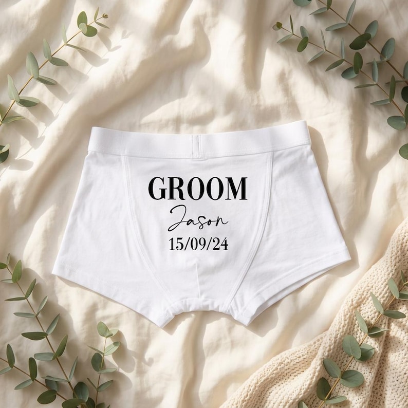 May include: White boxer briefs featuring black printed text that reads GROOM, followed by the name Jason in a script font, and the date 15/09/24. The garment has a wide elastic waistband and a central seam detail on the front panel.