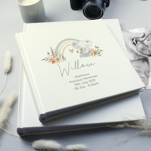 May include: A square white photo album featuring a printed illustration of a gray elephant, a muted rainbow, and floral accents. The text reads Willow, Matthews, Precious Memories, 15th July 2023, and 7lb 3oz - 9:26am in black lettering.