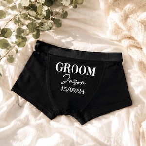 May include: Black boxer briefs featuring white printed text that reads GROOM, followed by the name Jason in a cursive script, and the date 15/09/24. The garment has a wide elastic waistband and visible stitching details on the front panel.