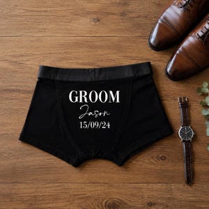 May include: Black boxer briefs featuring the white printed text GROOM, the name Jason in a script font, and the date 15/09/24. The garment has a solid black waistband and is displayed alongside a pair of brown leather dress shoes and a wristwatch.