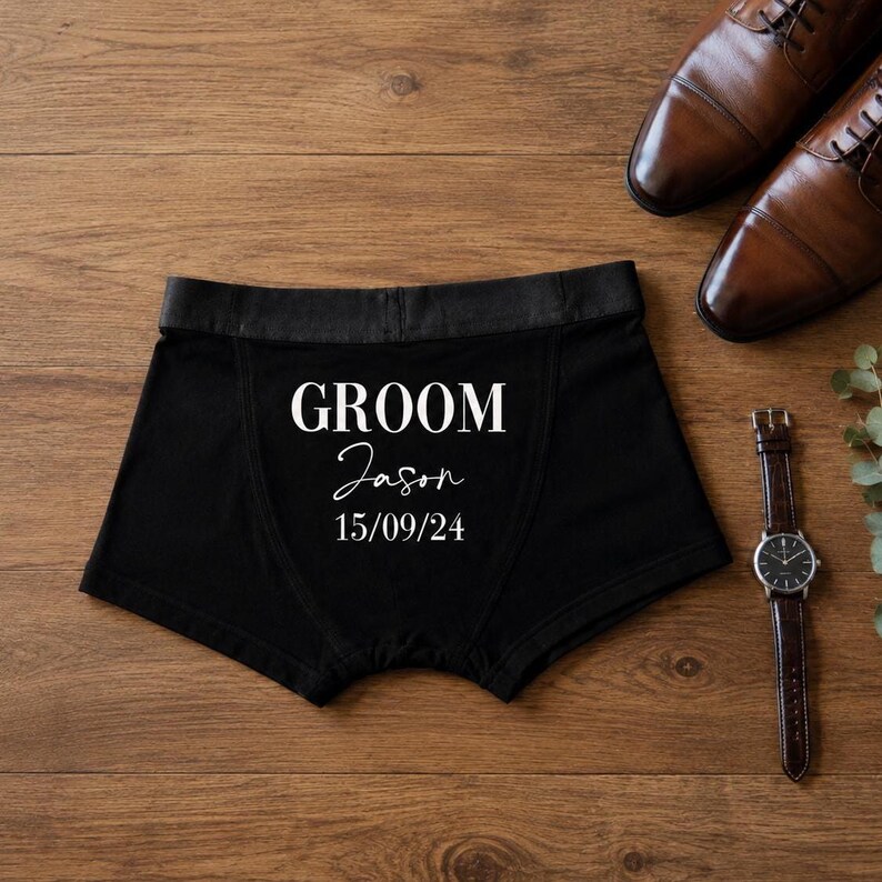 May include: Black boxer briefs featuring the white printed text GROOM, the name Jason in a script font, and the date 15/09/24. The garment has a solid black waistband and is displayed alongside a pair of brown leather dress shoes and a wristwatch.