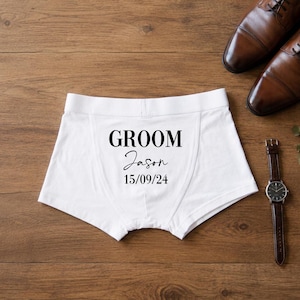 May include: White boxer briefs featuring the black printed text GROOM, the name Jason in a cursive script, and the date 15/09/24. The garment has a wide elastic waistband and a central seam, presented alongside a pair of brown leather dress shoes.