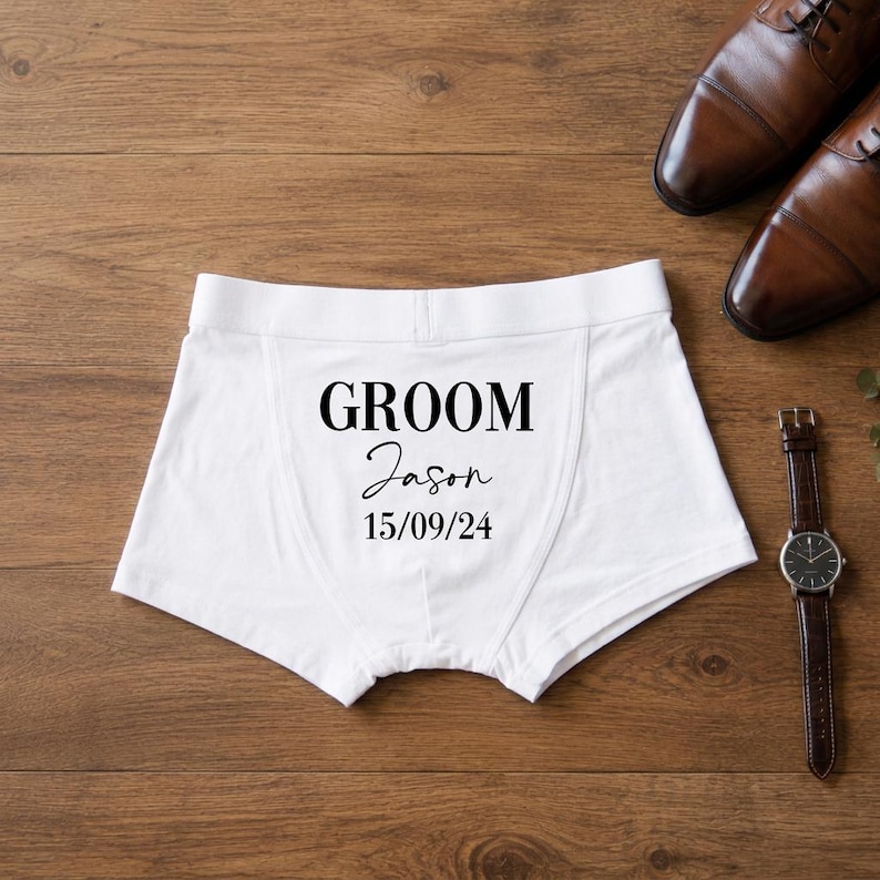 May include: White boxer briefs featuring the black printed text GROOM, the name Jason in a cursive script, and the date 15/09/24. The garment has a wide elastic waistband and a central seam, presented alongside a pair of brown leather dress shoes.