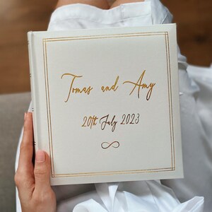 May include: A square, off-white photo album with a gold foil border. The cover features the gold text "Tomas and Amy", the date "20th July 2023", and an infinity symbol. The album has a clean, matte finish and a simple, elegant design.