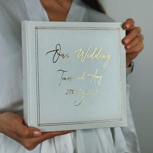 May include: A square, light grey wedding photo album featuring gold foil lettering that reads "Our Wedding", "Tomas and Amy", and "20th July 2023", with a gold infinity symbol and a thin gold border frame on the front cover.