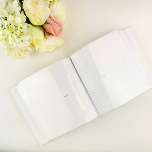 May include: An open photo album with white pages featuring clear plastic sleeves and lined memo areas next to each slot. The album is positioned next to a cluster of artificial cream, white, and soft pink peony flowers with green leaves.