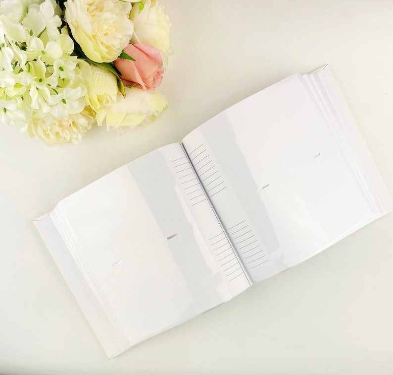 May include: An open photo album with white pages featuring clear plastic sleeves and lined memo areas next to each slot. The album is positioned next to a cluster of artificial cream, white, and soft pink peony flowers with green leaves.