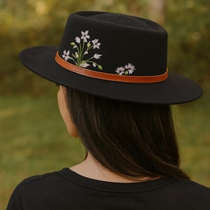 May include: Black fedora hat featuring a brown leather band and embroidered white floral detailing. The hat has a flat brim and a structured crown. A fashionable accessory for any ensemble.