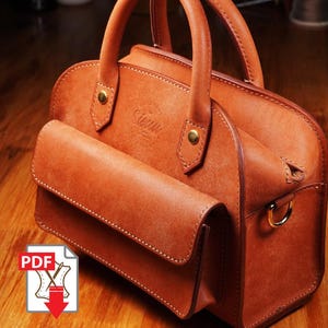 May include: A brown leather handbag with a front flap pocket and top handles. The bag features gold-colored hardware and visible stitching. The word "Sierra" is embossed on the front. The bag is sitting on a wooden surface.