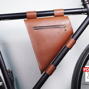 以下が含まれることがあります： 茶色の革製自転車フレームバッグ。ジッパー開閉と茶色の革ストラップ付き。バッグは黒い自転車フレームに取り付けられています。バッグは自転車のフレーム内に収まるように設計されています。