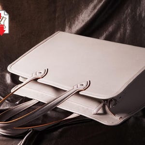 Peut inclure: Un sac à main en cuir gris clair de forme rectangulaire avec une poignée supérieure. Le sac est doté d'une fermeture à rabat et d'une fermeture éclair. La poignée est fixée avec du matériel métallique. Le sac est posé sur une surface sombre et texturée.