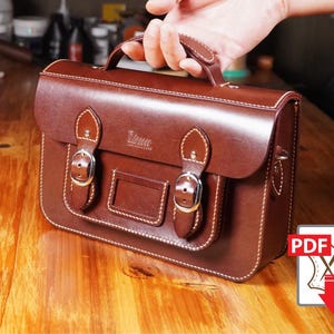 Pode incluir: Uma pasta de couro castanho com uma alça superior e dois fechos de fivela. A bolsa tem formato retangular com um pequeno bolso frontal e costuras brancas. A bolsa é segurada por uma mão e está sobre uma superfície de madeira.
