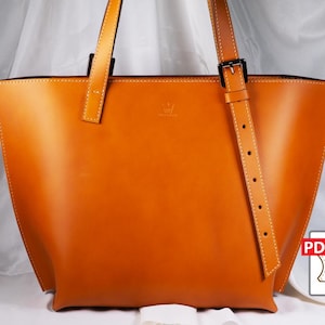 Peut inclure: Un grand sac fourre-tout en cuir brun orangé, de forme rectangulaire, avec une bandoulière. Le sac a deux poignées et une fermeture à boucle sur la bandoulière. Des coutures blanches ornent les bords. Une icône PDF est dans le coin inférieur droit.