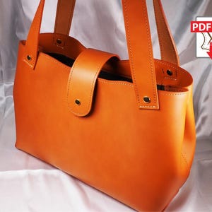 Könnte beinhalten: Orangefarbene Ledertasche mit rechteckiger Form und Klappenverschluss. Die Tasche hat zwei Griffe und goldfarbene Beschläge. Die Tasche ist aus glattem Leder gefertigt und scheint neu zu sein.