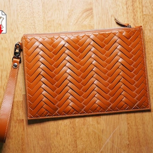 May include: A rectangular, woven leather clutch in a warm, caramel colour. The clutch features a zip closure and a matching wrist strap with a silver-tone clasp. The woven pattern creates a textured surface. The clutch is shown on a wooden surface.