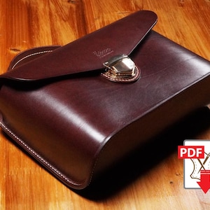 Puede incluir: Un bolso de cuero burdeos con un diseño curvo y cierre de solapa. El bolso presenta un cierre dorado y costuras blancas. El bolso está sobre una superficie de madera. Un icono PDF está en la esquina inferior derecha.