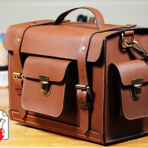 May include: A brown leather satchel with two front pockets, a top handle, and a shoulder strap. The bag has gold-coloured clasps and stitching details. The bag is a PDF download.