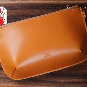 May include: A tan leather clutch bag with a zip and wrist strap. The bag has a minimalist design with a subtle embossed logo. The leather has a smooth, polished finish. The stitching is visible along the edges.