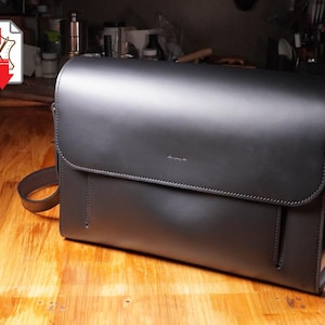 May include: A black leather messenger bag with a flap closure, shoulder strap, and a handle. The bag is rectangular and sits on a wooden surface. A PDF icon with a red arrow is in the upper left corner.