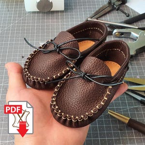 May include: A pair of brown leather baby moccasins with a textured finish. The shoes have a decorative stitched edge and black leather laces. The image includes a PDF icon with a sewing machine and a downward arrow.