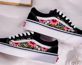 Peut inclure: Des baskets en toile noire avec des semelles blanches et un motif de broderie florale. La broderie présente des fleurs roses, rouges, vertes et blanches. Le mot "Helena" est brodé sur le côté de la chaussure.