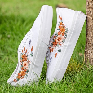 May include: Pair of white canvas sneakers with embroidered orange and green floral designs. The shoes have a classic low-top style with white soles and laces. The sneakers are leaning against a tree trunk in a grassy area.