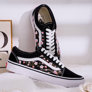 Könnte beinhalten: Schwarze Canvas-Sneaker mit weißen Sohlen und Schnürsenkeln. Die Sneaker haben ein Blumenstickerei-Design mit rosa und weißen Blumen und grünen Blättern. Der ikonische Seitenstreifen ist weiß. Die Schuhe werden auf einer weißen Oberfläche präsentiert.