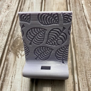 Op de afbeelding: Een lichtpaarse 3D-geprinte telefoonstandaard met een patroon van gestileerde bladeren. De standaard heeft een uitsparing voor een telefoon om in te rusten.