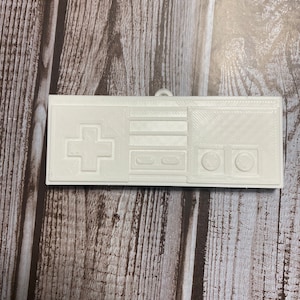 May include: White plastic ornament shaped like a vintage video game controller. The controller has a plus sign button, a directional pad, and four round buttons.