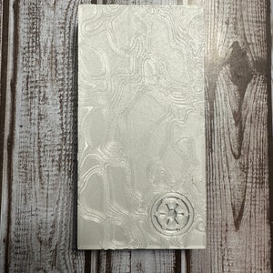 May include: A silver notebook with a textured surface and a raised, silver Imperial symbol from Star Wars.