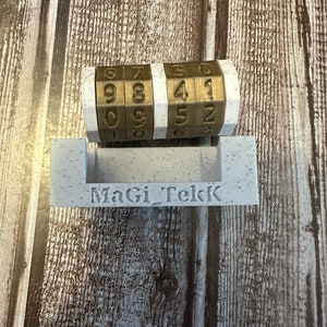 May include: A gold-colored combination lock with three rotating dials, each with numbers 0-9. The lock is mounted on a white plastic base with the text "MaGi_Tekk" printed on it.