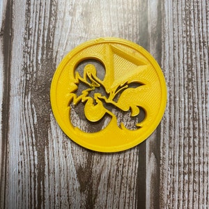 Op de afbeelding: Een gele plastic koekjesvormer in de vorm van een Dungeons and Dragons-symbool, een 20-zijdige dobbelsteen met een draak.