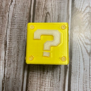May include: A yellow 3D printed question mark block with a white question mark. The block has four small white circles on the sides.