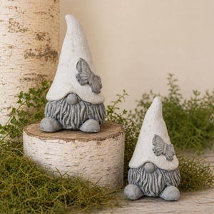 May include: Two gray and white garden gnomes with butterfly accents. The gnomes have long, pointed hats and textured beards. They are sitting on a birch log, surrounded by greenery, creating a whimsical outdoor decor theme.