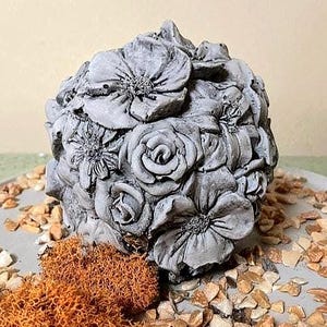 May include: A spherical concrete sculpture featuring a dense arrangement of sculpted flowers including roses and daisies. The surface has a matte gray finish with detailed petal textures and deep indentations between the individual floral elements.