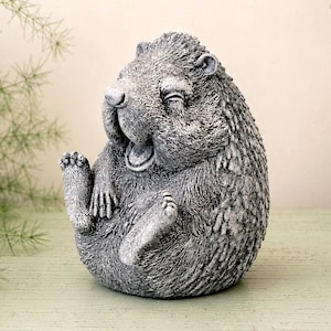 May include: A gray, textured stone-like figurine of a hedgehog sitting in an upright position with its mouth open as if laughing. The hedgehog has detailed fur textures, visible paws, and closed eyes, capturing a playful expression in a compact form.