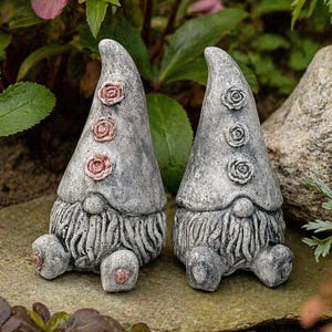 May include: Two gray garden gnomes with pointed hats and long beards. Each gnome has three rose-shaped embellishments on the hat and sits with legs extended. The gnomes are on a stone surface, surrounded by greenery and pink flowers.