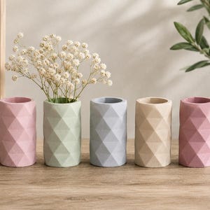 May include: Five cylindrical vases in pastel colors: pink, green, gray, beige, and pink. Each vase has a geometric diamond pattern. One vase holds white flowers. The vases are on a wooden surface.