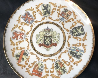 May include: A white ceramic plate with a gold rim and a gold design. The plate features the coats of arms of the provinces of Belgium, with the text "Souvenir de Belgique Gedenkenis van Belgie" in gold lettering.