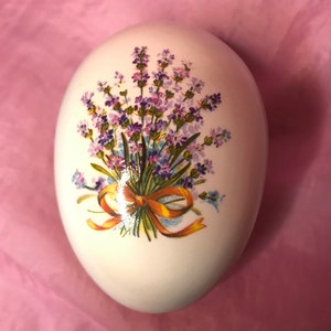 May include: White ceramic egg decorated with a floral design of lavender flowers and a yellow ribbon.
