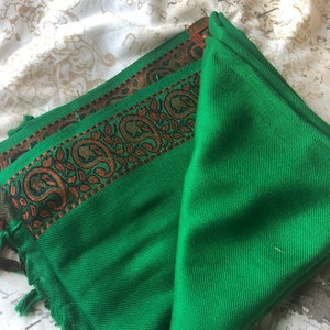 May include: A folded green scarf with a decorative border featuring a paisley design in shades of brown and orange. The scarf has a fringe of green tassels along the edge. The scarf is made of a soft material.