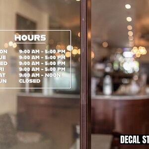 May include: A white decal with black text listing business hours. The decal is on a glass door with a blurred background of a cafe interior. The text reads: HOURS, MON 9:00 AM - 5:00 PM, TUE 9:00 AM - 5:00 PM, WED 9:00 AM - 5:00 PM, FRI 9:00 AM - 5:00 PM, SAT 9:00 AM - NOON, SUN CLOSED.