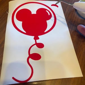 May include: A red Mickey Mouse balloon vinyl decal. The balloon design features the iconic mouse ears within a circular outline, with a curly tail. The decal is on a white backing, with a tool removing a piece of the vinyl.