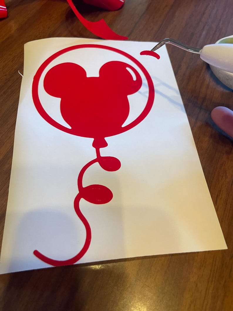 May include: A red Mickey Mouse balloon vinyl decal. The balloon design features the iconic mouse ears within a circular outline, with a curly tail. The decal is on a white backing, with a tool removing a piece of the vinyl.