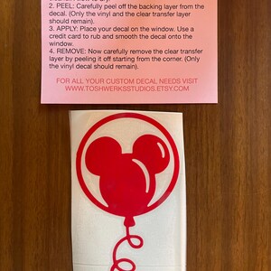 May include: A red vinyl decal featuring a Mickey Mouse balloon design. The decal is on a white backing, with instructions on a pink paper above. The instructions detail how to apply the decal to a window or smooth surface.